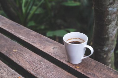 Yeşil arka planda, ahşap bankta bir fincan sıcak kahvenin yakın plan görüntüsü.