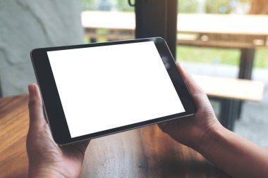 Ahşap masanın üzerinde boş beyaz masaüstü ekranı olan siyah tableti tutan ellerin görüntüsü