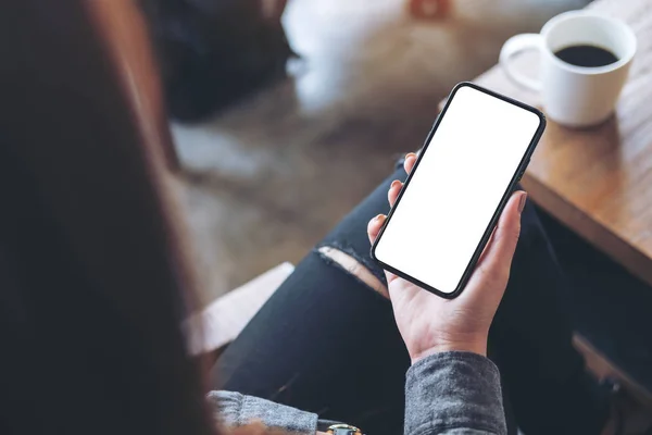 Kafede bacakları çapraz oturan, elinde siyah cep telefonu olan beyaz ekranlı bir kadının görüntüsü.