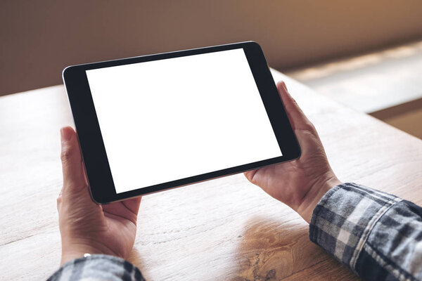 Mockup image of hands holding black tablet pc with blank white desktop screen on wooden table