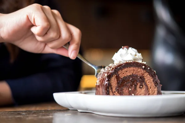 A woman's hand cutting chocolate roll cake and whipped cream on the top with fork in vintage wooden cafe