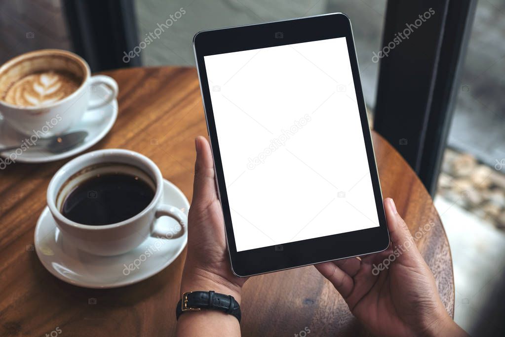 Mockup image of hands holding black tablet pc with white blank screen and coffee cups on table 