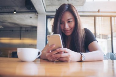 Portre resim tutarak ve modern loft Cafe ahşap masa üzerinde beyaz kahve fincanı ile akıllı telefon kullanarak gülen yüz ile güzel bir Asya kadının