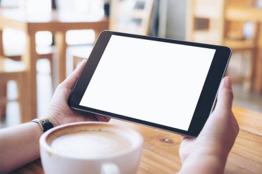 Mockup görüntü iş kadının elleri boş beyaz ekran ve ahşap masa üzerinde kahve fincanı ile siyah tablet pc kafe arka planda tutan