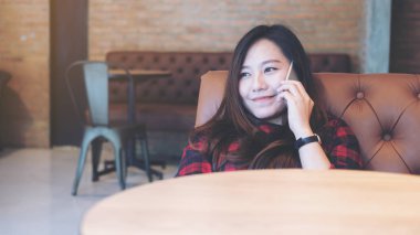 Bir surat güzel Asyalı kadın kahverengi kanepede yatan, ahşap masa ön plan ile modern kafede akıllı telefonda konuşurken ve kullanma