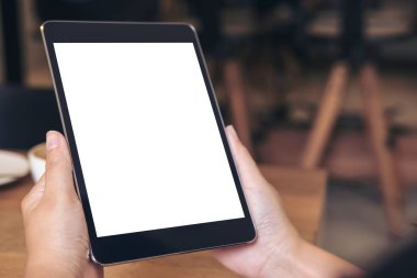 Kafe 'deki ahşap masada kahve fincanı ve beyaz ekranlı siyah tableti tutan ellerin görüntüsü.