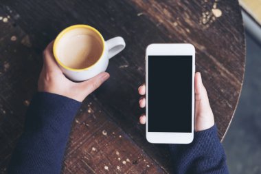 Kadının görüntüsünü holding beyaz cep telefonu ile boş siyah perde ve bir kahve fincanı vintage Cafe ahşap masa üzerinde eller mockup üstten görünüm