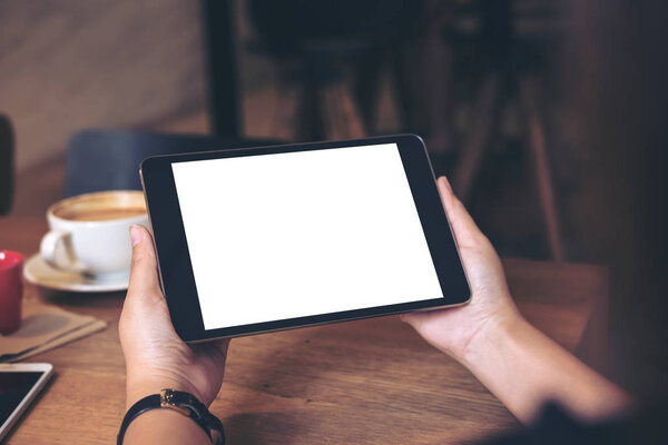Изображение рук, держащих черный планшет с чистым белым экраном с кофейной чашкой и мобильным телефоном на деревянном столе в кафе
