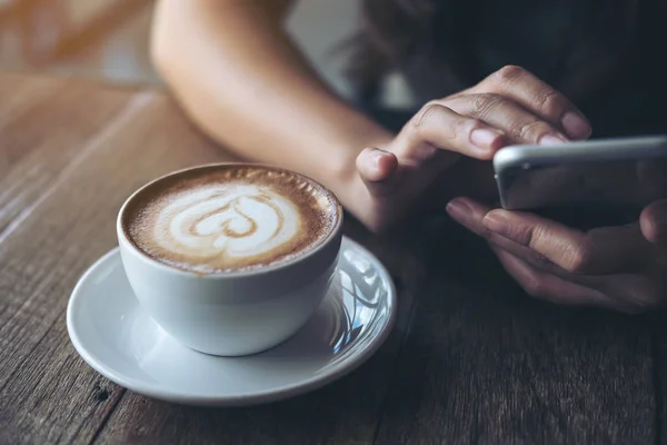 Holding, kullanma ve ahşap masa vintage cafe latte kahve Kupası ile akıllı telefon gösteren bir kadının görüntüsünü closeup eller
