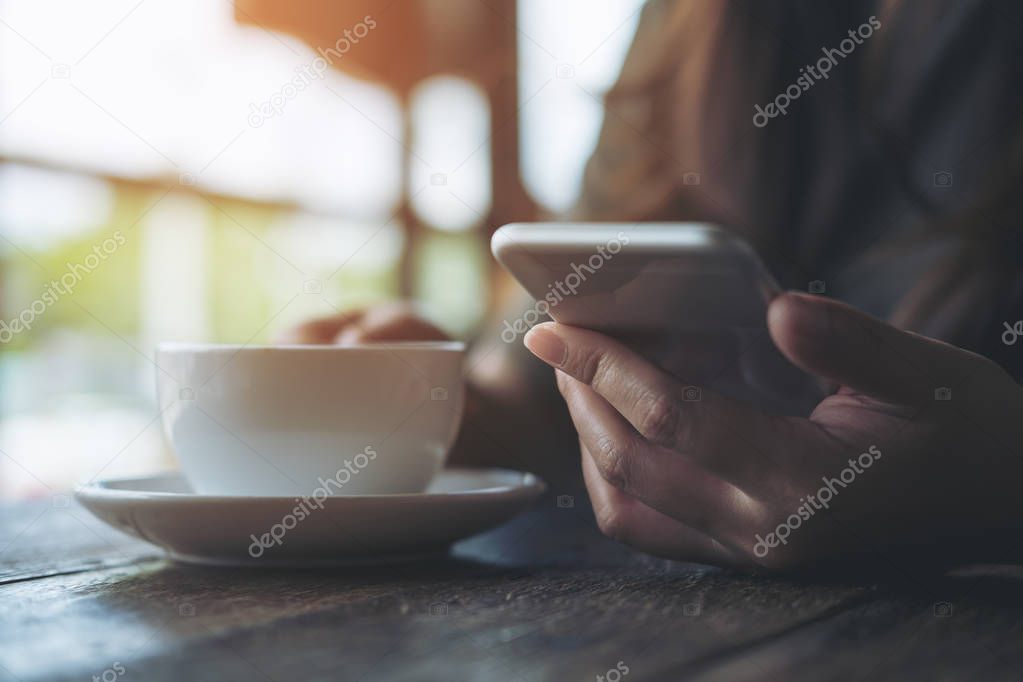 Closeup image of a woman using and looking at smart phone while holding a white coffee cup in modern cafe