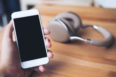 Bir erkek eli boş siyah perde ve kahverengi kulaklık ile beyaz cep telefonu kafede vintage ahşap masa üzerinde tutarak mockup görüntü
