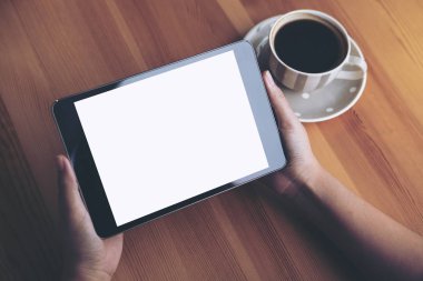 Mockup görüntü iş kadının elleri boş beyaz ekran ve ahşap masa üzerinde kahve fincanı ile siyah tablet pc kafe arka planda tutan