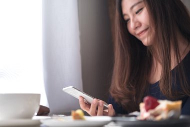 Bir surat görüntüsünü closeup holding, kullanma ve vintage Cafe ahşap masa üzerinde beyaz kahve Kupası ve tatlı tabağı ile akıllı telefon arıyorum güzel Asyalı kadın