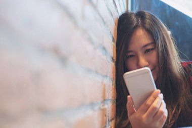 Bir surat closeup görüntüsünü kullanarak ve modern café'akıllı telefon bakarak tuğla duvara yaslanmış güzel Asyalı kadın
