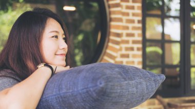 Güzel Asyalı kadın oturup üzerinde mavi bir yastık ile mutlu hissediyor ve kafede rahatla ellerini yukarıda çene ile tuğla duvar ve doğa arka plan