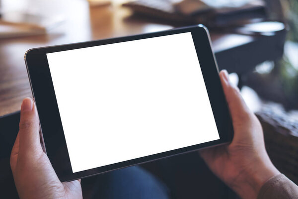 Mockup image of hands holding black tablet pc with white blank screen on wooden table background in cafe