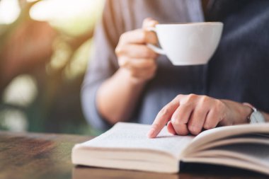 Ahşap masada kahve içerken kitap okuyup işaret eden bir kadının yakın plan resmi.