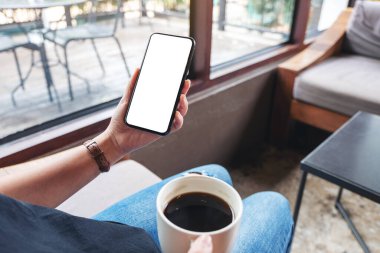 Kafede kahve içerken elinde boş masaüstü ekran olan siyah bir cep telefonu tutan kadının görüntüsü.