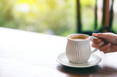 Yakından bakınca, beyaz bir fincan sıcak kahveyi ahşap masada tutan bir elin görüntüsü.