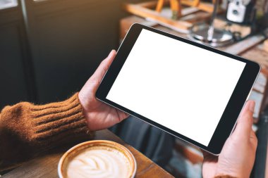 Siyah tabletli, beyaz ekranlı ve ahşap masada kahve fincanı olan ellerin görüntüsü. 