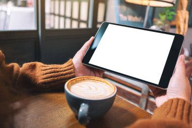 Siyah tabletli, beyaz ekranlı ve ahşap masada kahve fincanı olan ellerin görüntüsü. 