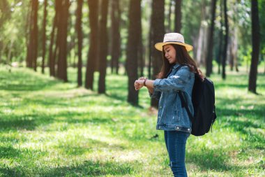 Güzel bir çam ormanındaki saate bakan şapkalı ve sırt çantalı Asyalı bir kadın gezgin.