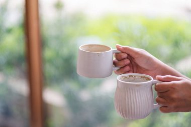 Yakın planda iki beyaz kahve bardağı tutan ve arka planda bulanıklık olan ellerin görüntüsü.