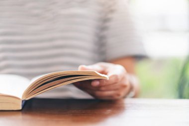 Ahşap masada elinde kitap tutan ve okuyan bir kadının yakın plan resmi.