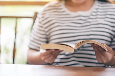 Ahşap masada elinde kitap tutan ve okuyan bir kadının yakın plan resmi.