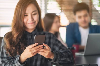 Güzel bir Asyalı kadın arka planda dizüstü bilgisayarla çalışan meslektaşlarıyla cep telefonuna bakıyor ve kullanıyor.