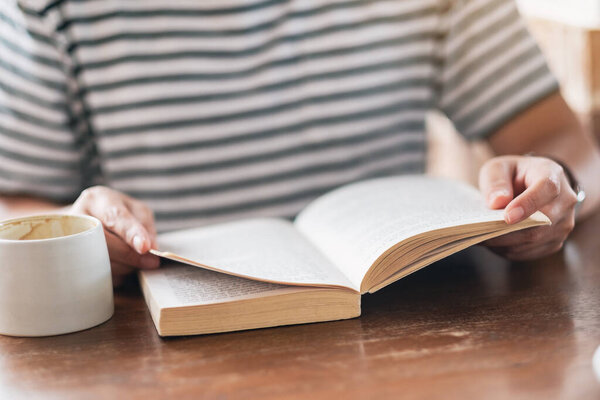 Closeup image of a woman holding and reading a book with coffee cup on wooden table