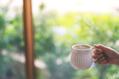 Yakın plan, arka planı bulanık beyaz bir fincan kahve tutan bir elin görüntüsü.