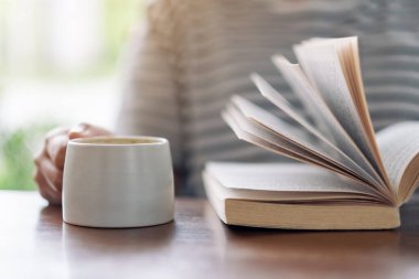 Tahta masada kahve içerken elinde kitap tutan ve okuyan bir kadının yakın plan resmi.