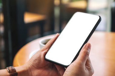 Masanın üzerinde kahve fincanı ve boş masaüstü ekranı olan siyah cep telefonunu tutan ellerin görüntüsü.