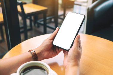 Masanın üzerinde kahve fincanı ve boş masaüstü ekranı olan siyah cep telefonunu tutan ellerin görüntüsü.