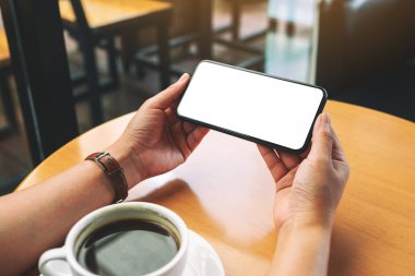 Masanın üzerinde kahve fincanı ve boş masaüstü ekranı olan siyah cep telefonunu tutan ellerin görüntüsü.