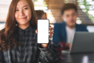 Siyah cep telefonunu elinde tutan ve arka planda çalışan meslektaşlarıyla ekranı boş gösteren bir kadının görüntüsü.