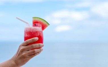 Yakından bakınca, deniz kenarında mavi gökyüzü arkaplanlı bir bardak karpuz kokteyli tutan bir elin görüntüsü.