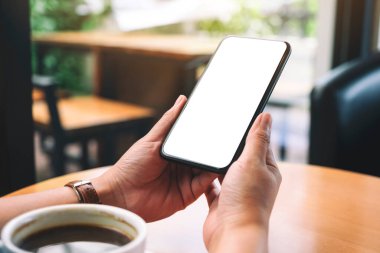 Masanın üzerinde kahve fincanı ve boş masaüstü ekranı olan siyah cep telefonunu tutan ellerin görüntüsü.