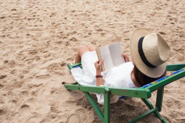 Güzel bir kadın uzanıp plaj sandalyesinde kitap okuyor ve rahatlıyor.