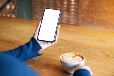 Kafedeki ahşap masada kahve içerken elinde beyaz bir cep telefonu olan bir kadının görüntüsü.