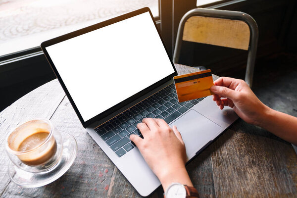 Изображение руки, держащей кредитную карту во время использования и ввода на ноутбуке с чистым белым экраном и чашкой кофе на деревянном столе