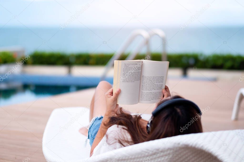 A woman listening to music with headphone and reading a book while lying down by swimming pool