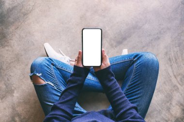 Üst görünümde, yerde otururken elinde beyaz ekranlı siyah bir cep telefonu tutan bir kadının görüntüsü var.