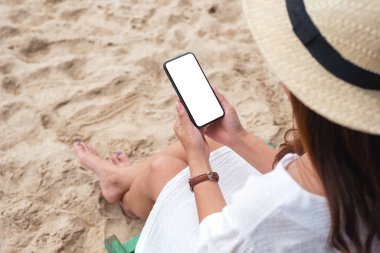 Plaj sandalyesinde otururken elinde boş masaüstü ekran olan siyah bir cep telefonu tutan bir kadının görüntüsü. 