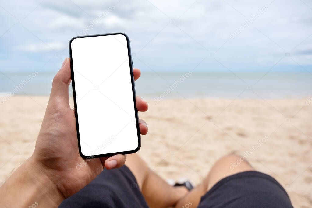 Mockup image of a man's hand holding black mobile phone with blank desktop screen while laying down on beach chair