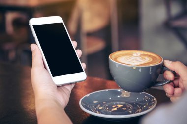 Modern kafede kahve içerken beyaz cep telefonunu boş ekran ile tutan ellerin görüntüsü.