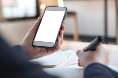 Ofisteki deftere yazarken beyaz cep telefonunu beyaz ekranla tutan ellerin görüntüsü