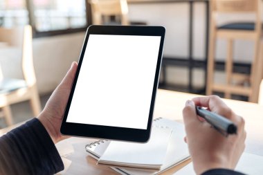 Ofisteki not defterlerine yazarken boş beyaz masaüstü ekran ile siyah tablet pc 'yi tutan ve kullanan ellerin görüntüsünü taklit et