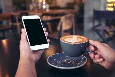 Modern kafede kahve içerken beyaz cep telefonunu boş ekran ile tutan ellerin görüntüsü.